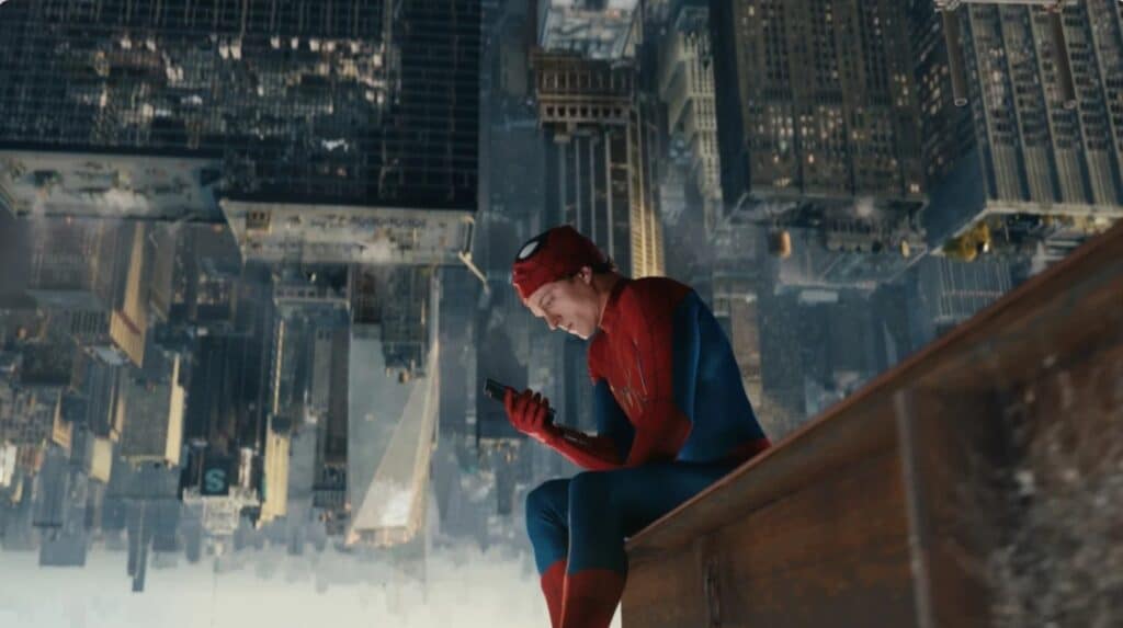 Tom Holland’s Spider-Man sits on a high ledge above an inverted New York skyline, looking at a phone — a still that captures the film’s isolation. Alt text: Spider-Man in the red and blue suit sitting on a wooden rooftop beam, mask pulled up, staring at a smartphone while the Manhattan skyline appears upside down behind him in Spider-Man Brand New Day.
